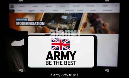 Person holding cellphone with logo of United Kingdom ground force British Army in front of webpage. Focus on phone display. Stock Photo
