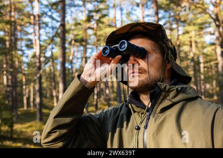 man in camouflage clothing looking through binoculars in forest. park monitoring, bird watching Stock Photo