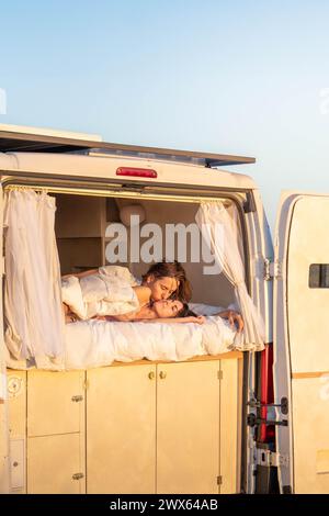Young couple kissing reclining on a bed in the back of a camper van, gazing out at the sunrise with a sense of tranquility. Stock Photo
