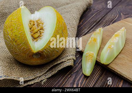 Close-up of a cut melon lying on a wooden table with a burlap bedding, next to a cutting board with fruit slices Stock Photo