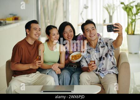 Group of smiling friends sitting on couch at home and taking selfie Stock Photo