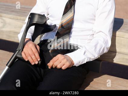 A man in formal wear, holding crutches, sits on a bench Stock Photo