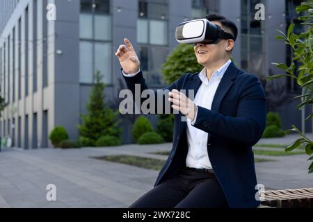 Smiling young man sitting outside office center on bench wearing virtual glasses, gesturing with hands and working remotely, having fun on work break. Stock Photo