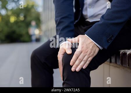Close-up photo of young man's hands in a business suit holding the knee of the leg feeling severe pain. Stock Photo