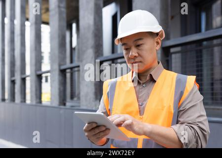 Asian young man, construction worker, foreman, company owner stands outside in hard hat and vest and concentrates on tablet. Stock Photo
