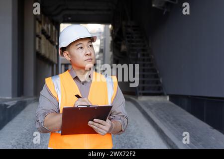 Asian young man builder, expert and inspector inspects and records work progress. Standing in a hard hat and vest, looking around and writing documents. Stock Photo