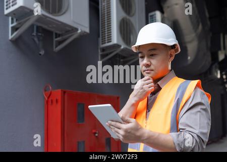 Close-up photo of a thoughtful and serious young Asian man, a factory worker, a construction worker and an engineer standing outside in a hard hat and vest, using a tablet. Stock Photo