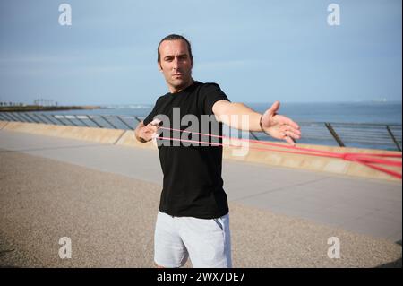 Young athlete doing arms, biceps and shoulder exercises with resistance fitness rubber bands on the beach outdoors. Handsome confident determined athl Stock Photo