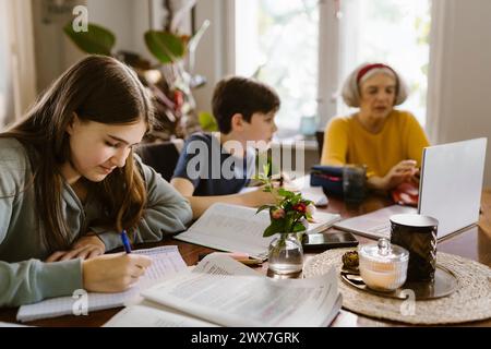 Girl doing homework while sitting with brother and grandmother at home Stock Photo