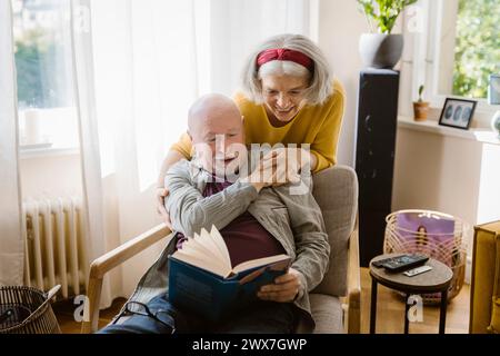 Happy senior woman with arm around man reading book while sitting on chair at home Stock Photo