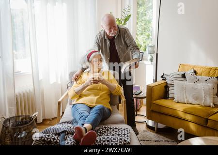 Senior man showing book to woman sitting on chair and holding cup at home Stock Photo