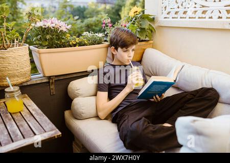 Boy drinking juice and reading book while reclining on sofa in balcony Stock Photo