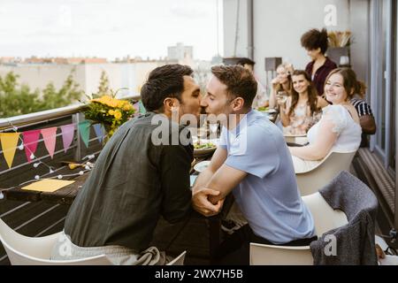 Affectionate gay couple kissing while sitting with friends at dinner party in balcony Stock Photo