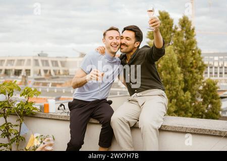 Carefree gay couple toasting drinks while sitting on wall during party in balcony Stock Photo