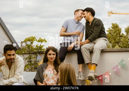 Happy gay couple sitting on wall with friends talking with each other at party in balcony Stock Photo