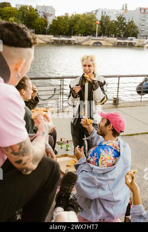 Happy non-binary person having pizza with friends near promenade in city Stock Photo
