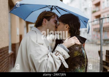 Side view of affectionate gay couple kissing under umbrella at street Stock Photo