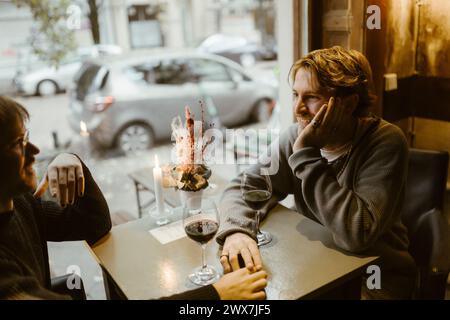 Smiling gay man leaning on elbow while talking with boyfriend sitting at restaurant Stock Photo