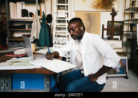 Businessman writing in diary while sitting with laptop on table at workshop Stock Photo
