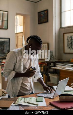 Male entrepreneur with disposable cup using laptop while standing at workshop Stock Photo