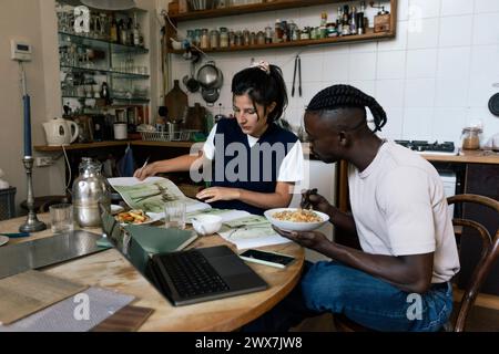 Male designer having lunch while female colleague examining blueprint sitting in home office Stock Photo