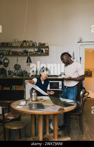 Female architect analyzing blueprint by male colleague having lunch in home office Stock Photo
