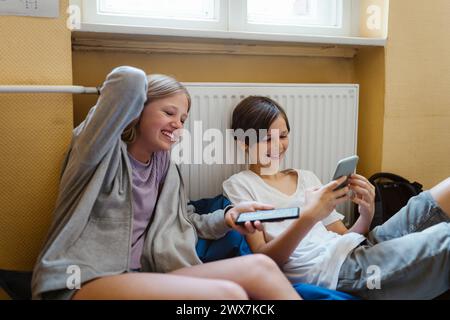 Happy male and female students using smart phone while sitting near heater at school Stock Photo