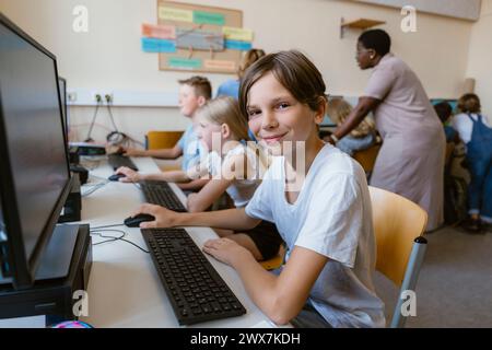 Portrait of smiling male pupil sitting with friends at desk in computer classroom Stock Photo