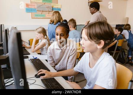 Smiling girl sitting with male friend using computer while sitting in classroom at school Stock Photo