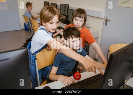 Curious girl pointing at computer near boy and female friend with disability in classroom Stock Photo