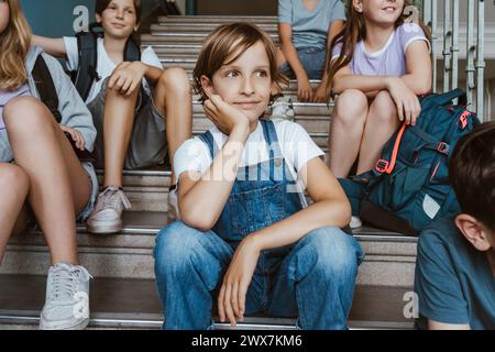 Thoughtful girl leaning on elbow while sitting on staircase with friends in school Stock Photo