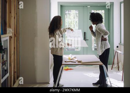 Side view of client and female architect comparing blueprints while standing near table at under construction apartment Stock Photo