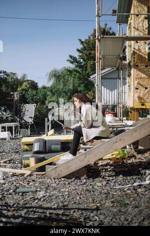 Side view of female architect talking on smart phone while sitting on steps at construction site Stock Photo
