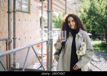 Portrait of smiling female architect holding blueprints while standing near under construction apartment Stock Photo