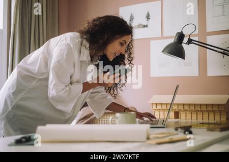 Smiling female construction freelancer talking through speaker phone and using laptop while working at office Stock Photo