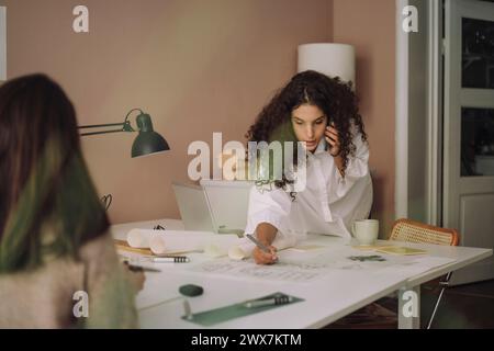 Female architect preparing blueprint while talking on smart phone at office Stock Photo