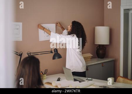Side view of female architect sticking blueprint on wall while working at office Stock Photo