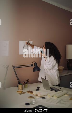 Busy female architect talking through speaker phone while examining blueprint at office Stock Photo
