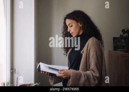 Female architect with curly hair reading book at home Stock Photo