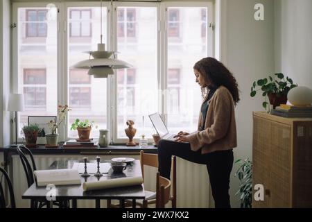 Side view of female construction engineer using laptop while standing with one leg on chair near table at home Stock Photo