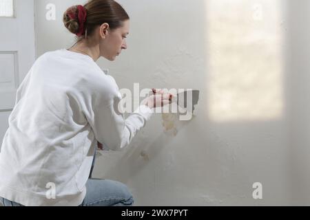 Saltpeter on the wall problem. Woman is using a scraper to scrape and remove all loose paint and plaster that is in poor condition, until a firm surfa Stock Photo