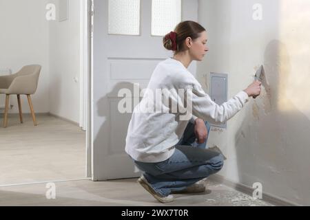 Saltpeter on the wall problem. Woman is using a scraper to scrape and remove all loose paint and plaster that is in poor condition, until a firm surfa Stock Photo