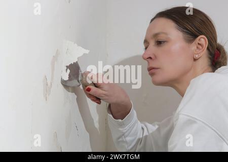 Saltpeter on the wall problem. Woman is using a scraper to scrape and remove all loose paint and plaster that is in poor condition, until a firm surfa Stock Photo