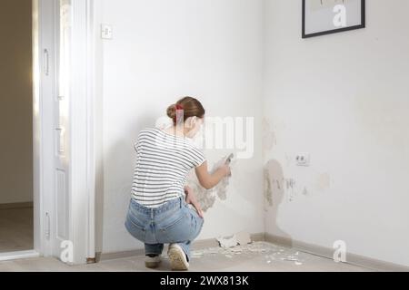 Saltpeter on the wall problem. Woman is using a scraper to scrape and remove all loose paint and plaster that is in poor condition, until a firm surfa Stock Photo