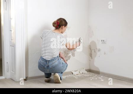 Saltpeter on the wall problem. Woman is using a scraper to scrape and remove all loose paint and plaster that is in poor condition, until a firm surfa Stock Photo