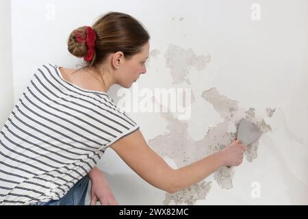 Saltpeter on the wall problem. Woman is using a scraper to scrape and remove all loose paint and plaster that is in poor condition, until a firm surfa Stock Photo