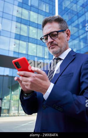 Business senior man using phone to work, standing outside of a corporate district. Stock Photo