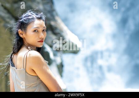 A young woman stands in front of a waterfall and turns to the camera Stock Photo