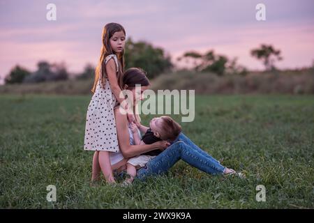 Mother embraces son and daughter, sitting on grassy field as twilight settles. Moment of tranquil affection, connection. Serene Family Time at Dusk. W Stock Photo