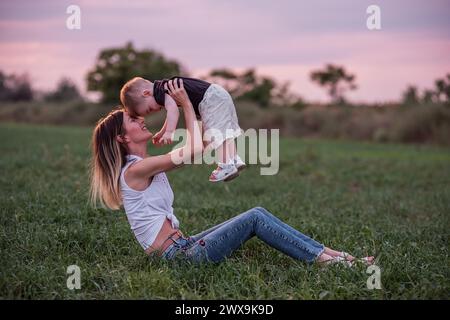 Smiling woman lifts, kiss little boy high in grassy field during serene sunset. Concept of pure joy, bonding, family connection. Mother and son walks Stock Photo